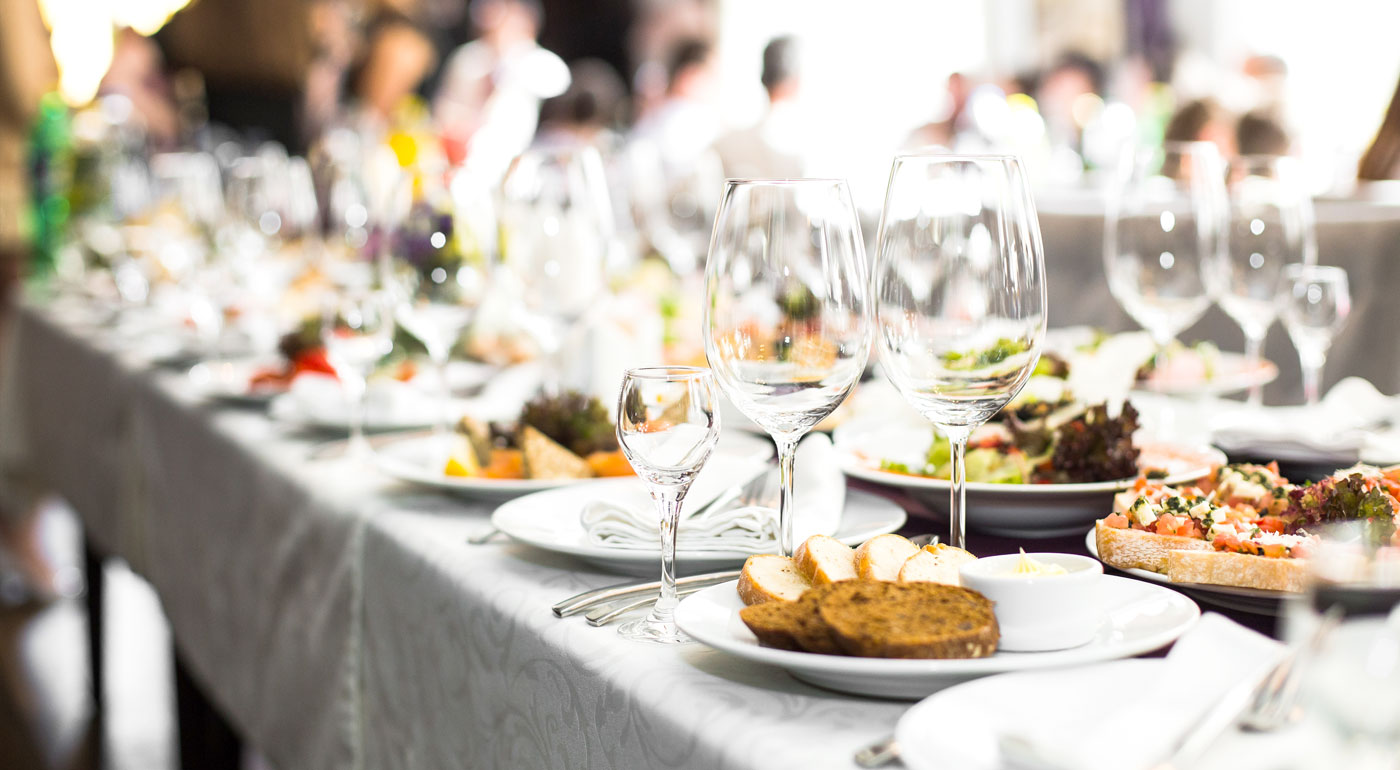 Nahaufnahme einer Langen Tafel mit Gästen im Hintergrund. Es ist viel Miet Geschirr und leckeres Catering Essen zu sehen.